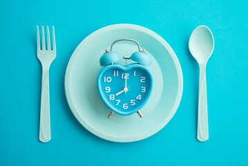 Flat lay of minimal style blue retro heart clock alarm on blue pastel disk, fork and spoon on blue background. IF (intermittent fasting), diet and weight loss, creative idea design, art concept.