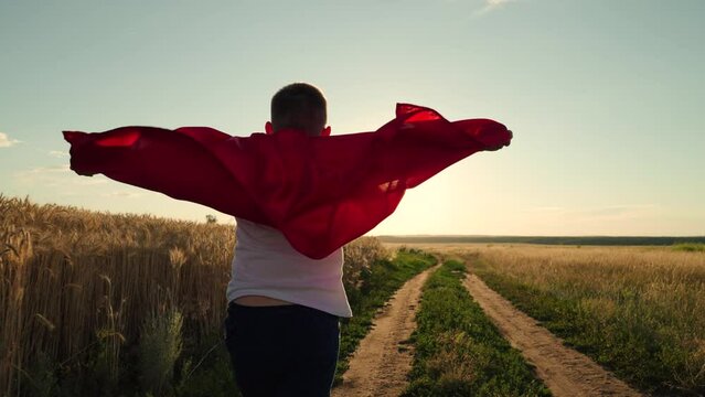 Child, superhero in red cape, nature, sky. Active Boy, child plays superhero in sun. Happy run of child in red raincoat at sunset park. Boy runs across green field with flowers, childhood dream to fly