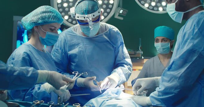 Diverse team of professional surgeons performs invasive surgery on patient in hospital. Nurse gives necessary tools to doctor. African-American assistant helps head surgeon apply sutures.