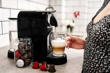 Girl with capsule coffee machine and cappuccino in transparent cup at kitchen. Woman preparing...