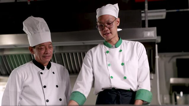 4k happy middle age man working together with junior chef in restaurant, head chef in unifrom train new worker how to cook or stir ingredients, good collaboration working lifestyle concept