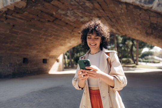 Young smiling woman wearing casual clothes holding smartphone using cellphone modern technology, looking at mobile, checking cell phone apps, texting, browsing internet while walking down the street.