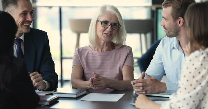 Positive confident senior business leader woman in glasses talking to younger colleagues on meeting, discussing project strategy with team, smiling, laughing, chatting. Company mentor coaching interns