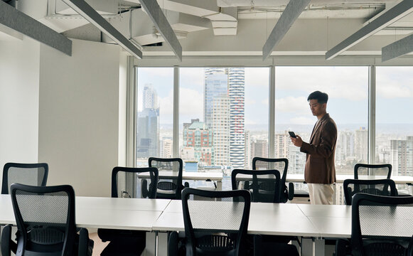 Young busy Asian business man using mobile cell phone tech standing in big modern office space. Professional Japanese businessman holding smartphone, working on cellphone corporate tech device.