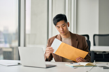 Young busy Asian professional business man manager opening envelope delivery receiving business mail letter holding paper documents bank statements, invoice file or bill sitting at desk in office.