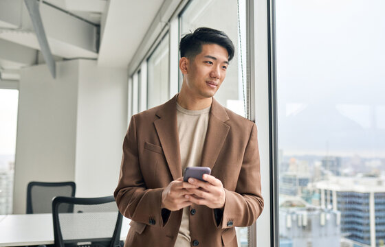 Asian young business man ceo leader holding phone looking at window standing in modern office. Japanese male professional entrepreneur using smartphone thinking of future leadership.
