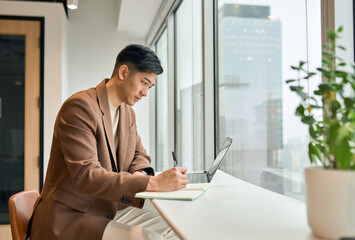 Young busy Asian business man manager working on laptop in office writing notes. Professional...