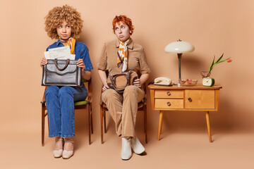 Thoughtful two retro women dressed in old fashionable clothes pose with bags sit next to each other have pensive expressions try to apply for job isolated over brown background. Back to 80s concept