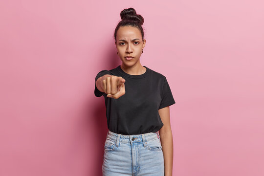 Serious dark haired woman with pleasant appearance points index finger at camera blames you for doing something wrong wears black t shirt and jeans says hey stop there poses against pink background.