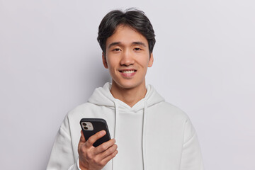 Horizontal shot of pleased Chinese make teenager uses smartphone sends messages pays for connection to app dressed in casual sweatshirt isolated over white background. Modern lifestyle concept