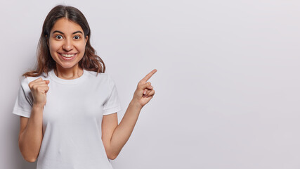 Studio shot of young Iranian woman with dark hair clenches fist points index finger on blank space...