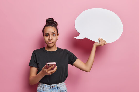 Latin woman holds speech bubble and smartphone invites you to imagine impactful words she may share raises eyebrows poses against pink background. Confused millennial girl with hair bun holds cloud