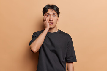 Surprised shocked dark haired Asian man keeps hand on cheek stares with stupefied expression at camera being terrified by something dressed in black casual t shirt isolated over brown background