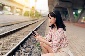 Fototapeta premium Asian young woman traveler with weaving basket using a mobile phone beside railway train station in Bangkok. Journey trip lifestyle, world travel explorer or Asia summer tourism concept.