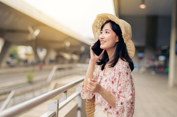 Asian young woman traveler with weaving basket using a mobile phone beside railway train station in Bangkok. Journey trip lifestyle, world travel explorer or Asia summer tourism concept.