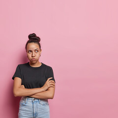 Vertical shot of offended Latin woman keeps arms folded looks unhappily aside being offended blows cheeks wears casual black t shirt and jeans isolated over pink background blank space for text