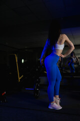 Fototapeta na wymiar A sports girl in blue leggings and a sexy body warms up in front of a mirror at a workout in a gym with exercise equipment 