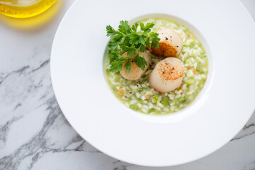 White plate with pea risotto and seared sea scallops, high angle view on a grey marble background,...