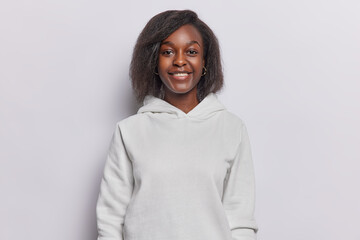 Portrait of good looking dark skinned woman with toothy smile dressed in casual sweatshirt looks directly at camera poses against white background. People emotions and face expressions concept