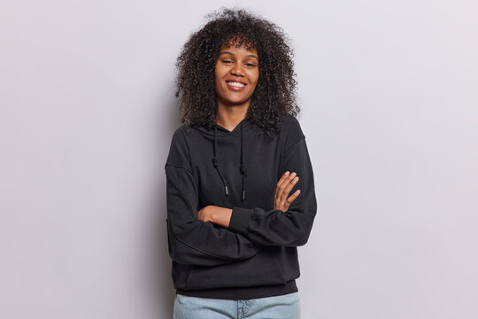 Indoor shot of cheerful self confident Afro woman keeps arms folded stands in assertive pose smiles pleasantly wears black sweatshirt jeans isolated on white background ready to give helpful advice