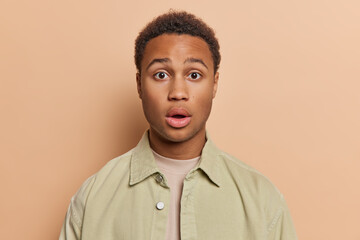 Portrait of handsome young Afro man with short curly hair keeps mouth widely opened holds breath from amazement wears stylish jacket isolated over brown background. People and reactions concept