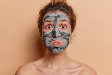 Surprised young female model engages in self-care applies rejuvenating face mask for flawless and youthful skin blows cheeks makes funny grimace stands topless indoor against brown background