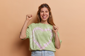 Triumphant young European woman raises clenched fists in celebration her beaming smile reveals sense of joy and accomplishment expresses pure happiness wears casual t shirt and jeans poses indoor