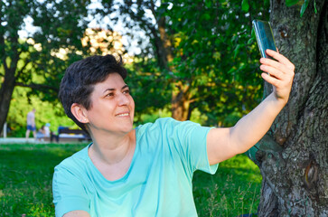 Smiling woman sitting on grass in park, takes selfie or communicating via video on smartphone