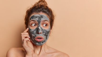 Stunned young woman luxuriates in spa treatment adornes her face with hydrating mask revitalizes complexion wants to have smooth skin stands shirtless against brown background copy space for text