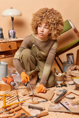 Fototapeta na wymiar Pensive curly female carpenter uses copig saw concentrated aside dressed in overalls and protective gloves does woodwork surrounded by variety of instruments produces wooden items in workroom