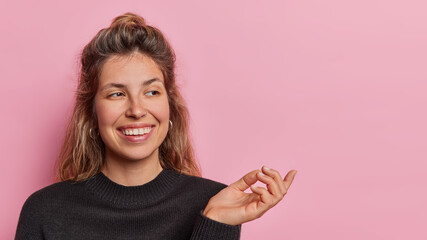 Horizontal shot of cheerful Caucasian woman with beaming pleasant smile on face keeps hand raised concentrated aside dressed casually stands against pink background copy space for your promotion