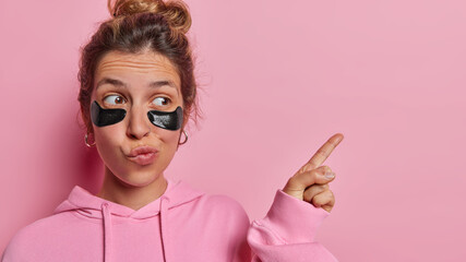 Beauty and wellness advertisement. Dark haired woman wears beauty black hydrogel patches showcases latest trend in skincare promots cosmetic product dressed in hoodie isolated over pink background.