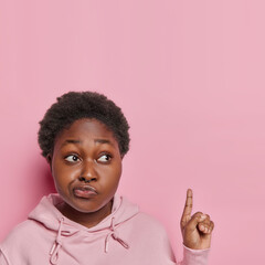 Marketing promotions advertising and retail concept. Young black woman points upwards gestures upwards her attention caught by captivating sale banner against pink background wears casual clothes