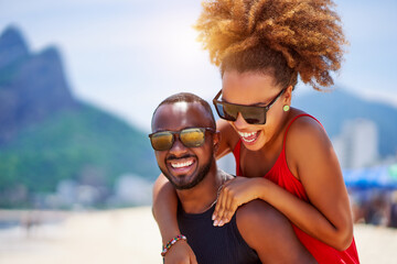 head shot portrait young black brazilian couple in piggyback position having fun in Ipanema beach Brazil - obrazy, fototapety, plakaty