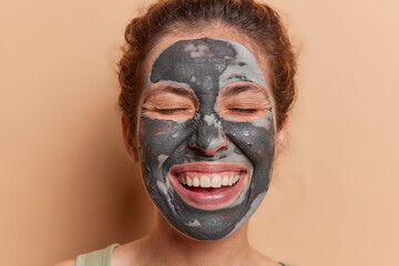 Graceful lady adorns flawless complexion with revitalizing face mask unlocking secrets to ageless beauty smiles broadly keeps eyes closed undergoes self care procedures isolated on brown wall