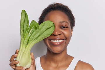 Headshot of dark skinned young woman holds bok choy over face smiles broadly being in good mood...