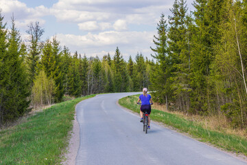 View of woman on bicycle moving along asphalt cycling road against backdrop of forest on both...