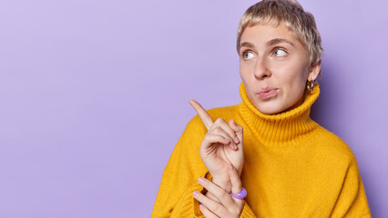 Serious blonde woman points index finger on copy space demonstrates copy space shows place for your advertisement dressed in warm yellow jumper isolated over purple background. Look at this.