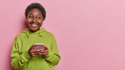 Joyful black plump woman holds mobile phone with radiant smile stays connected looks gladfully...