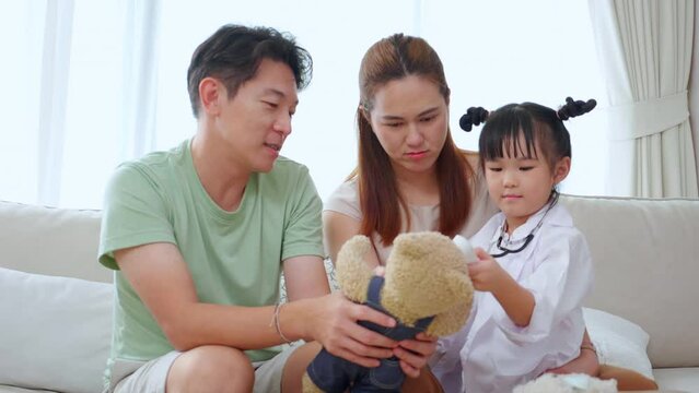 Happy family with daughter play doctor or nurse checkup and examining fever with teddy bear on sofa in living room at home, dream child, kid with imagination, lifestyles and medical concept.