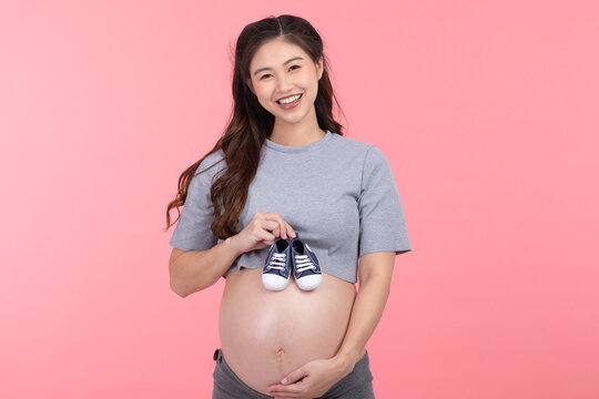 Asian pregnant woman holding baby sneakers for baby newborn isolated on pink background. Pregnant woman packing baby stuff ready for the maternity hospital. Pregnancy Prepare for Newborn Concept