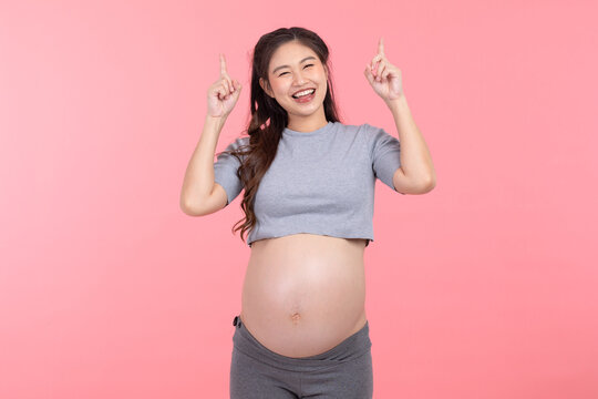 Happy Pregnant Woman standing smile stroking big belly with love and pointing up isolated on pink background,Pregnancy of young woman enjoy with future life,Motherhood and Pregnant Concept