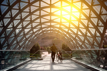 An elderly man with a bicycle walks along a beautiful bridge with a glass arch.