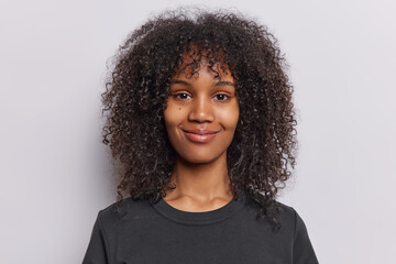 Portrait of pleased teenage girl with curly frizzy hair smiles gently expresses positivity and contentment looks pleased at camera dressed in casual black t shirt isolated over white background