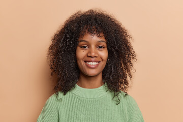 Portrait of lovely Afro woman with dark curly hair smiles pleasantly feels optimistic dressed in casual green knitted jumper poses against brown background. People and positive emotions concept