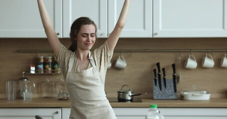 Cheerful excited energetic young woman in apron having fun in kitchen, dancing to music, singing song, smiling, laughing, moving at table, enjoying activity, leisure, motion