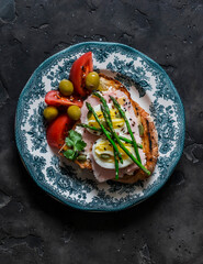 Sandwich with boiled egg, ham, asparagus and tomatoes, olives in a plate on a dark background, top view