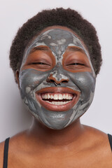 Cropped shot of overjoyed dark skinned woman has broad smile, keeps hands near face, applies clay mask, Afro hairstyle, enjoys softness of skin, isolated over pink background. Facial treatments
