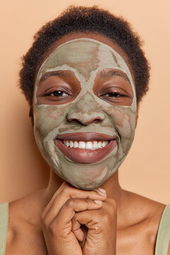 Vertical shot of cheerful dark haired woman smiles gently keeps hands under chin applies nourishing clay mask for skin rejuvenation undergoes beauty procedures poses against brown background