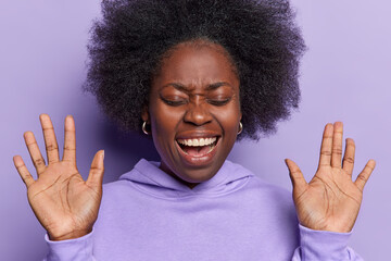 Headshot of emotional dark skinned woman with dark bushy hair keeps palms raised up exclaims loudly shows white teeth dressed in casual sweatshirt isolated over purple background. People and emotions